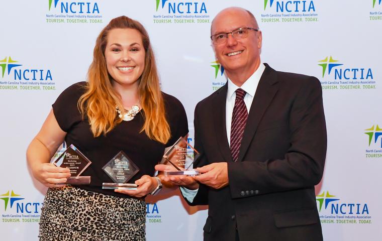 Two people stand, smiling, with 3 awards in-hand in front of a plain backdrop at a ceremony
