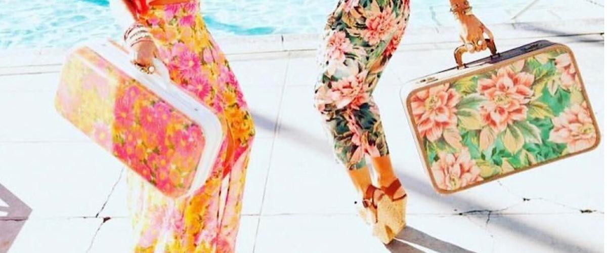 Image of two women's legs in colorful, floral pants carrying matching floral luggage.  They are standing next to a pool and look to be dancing.
