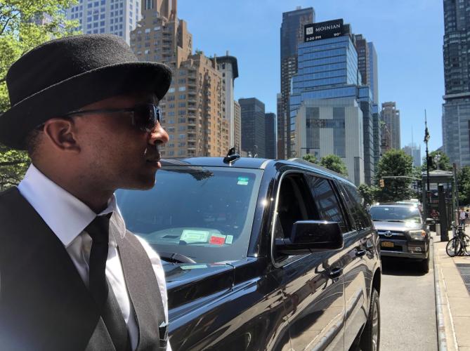 Upstate Living Transit driver wearing a top hat and suit, standing with limo with city skyline in the background.