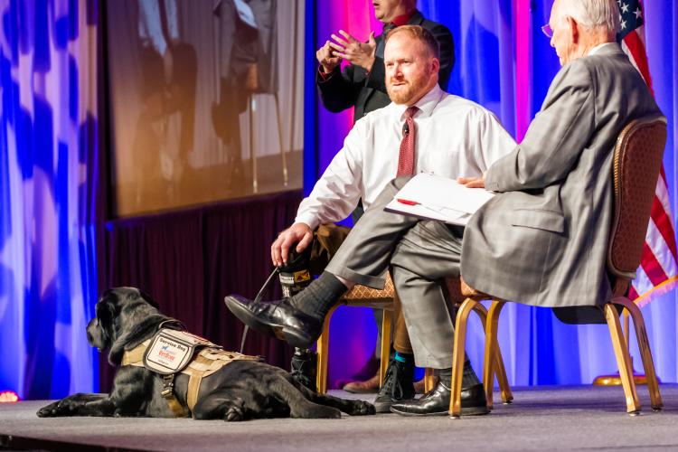 Two People speaking on a stage with a service dog laying down next to them