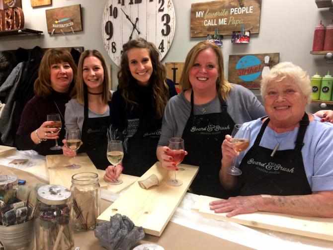 Five women smile for the camera with their wine in hand and boards in front of them ready to be painted