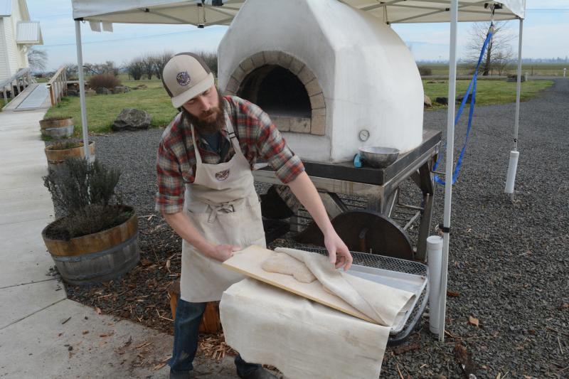 Baking Woodfired Bread at Camas Country Mill by Colin Morton Bearded man in a flannel shirt and an apron pulling pizza dough from under a cloth. He stands in front of an outdoor pizza oven at Camas Country Mill.