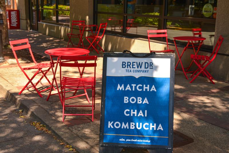 Brew Dr. Teahouse by Melanie Griffin A sign that says "Brew Dr. Tea Company- Matcha, Boba, Chai and Kombucha" sits in front of red outdoor dining tables with matching chairs.