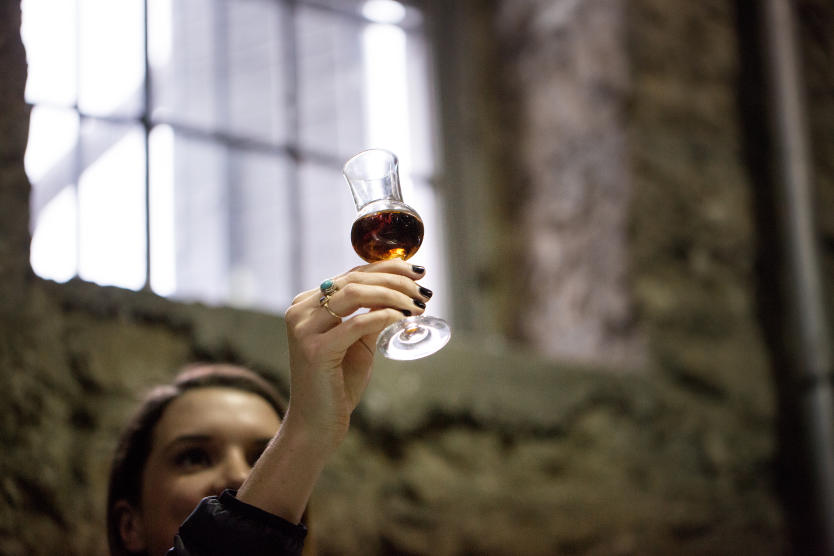 A woman holds a small tasting glass of bourbon up into the light from a window to view its color.