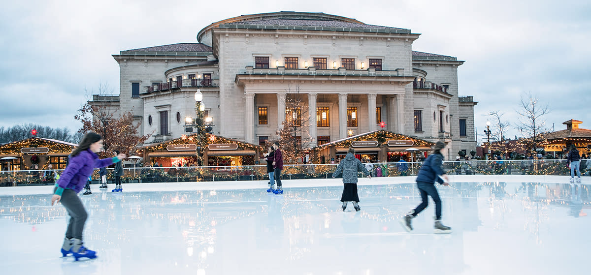 6 ice skaters on an ice rink in front of a building with holiday lights on it