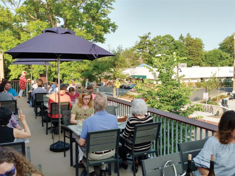 Groups of customers sit below umbrellas at the tables. The tables are on the restaurant's rooftop which overlooks the street.