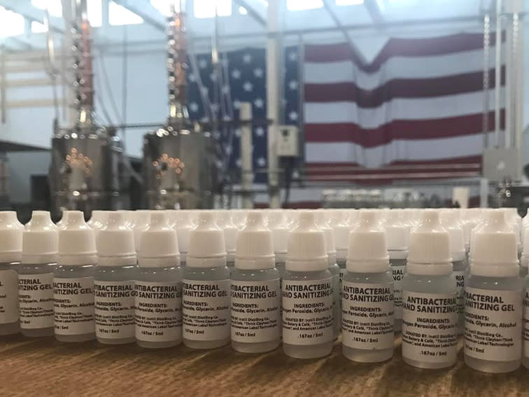 Bottles of hand sanitizer lined up on a table with a rum still and American flag in the background