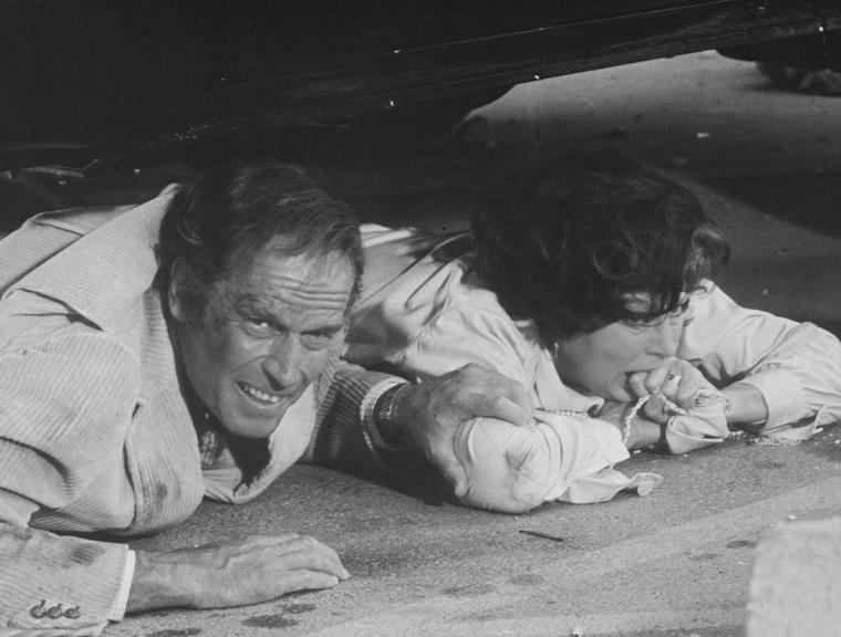 Ava Gardner and her Earthquake co-star lay under a car in this behind the scenes black and white photo.