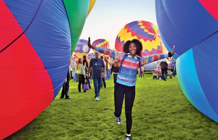 A girl stands between two hot air balloons with her arms stretched outrl