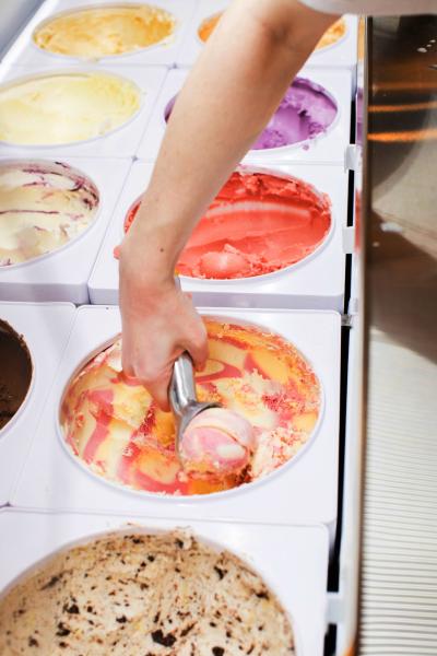 Hand scooping out of one of several large, colorful tubs of ice cream
