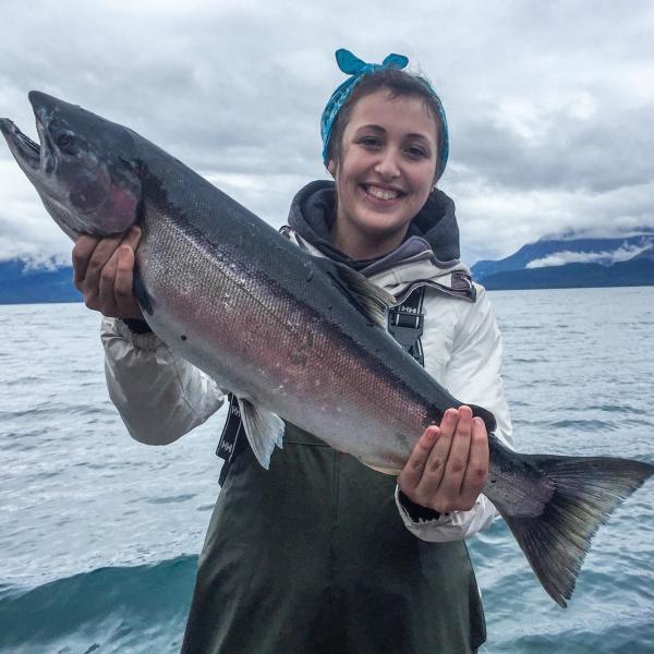 a woman holds up a large silver salmon; ocean and mountains in background