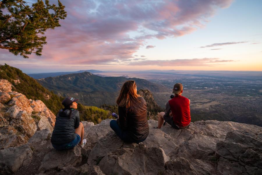 A view looking to the West at Sandia Crest with three people sitting on rocks in the foreground