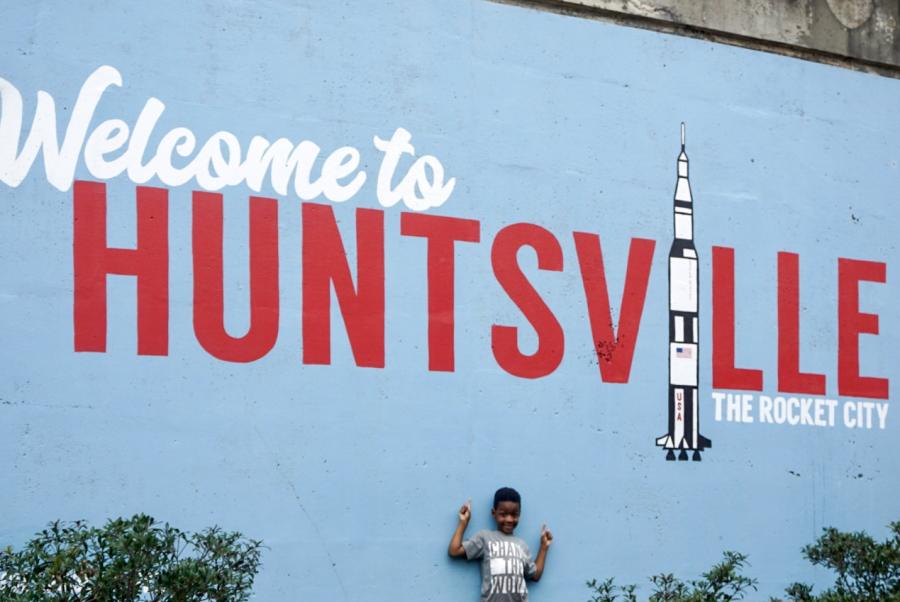 A child wearing a shirt that says "Change The World" poses in front of the Welcome to Huntsville mural.
