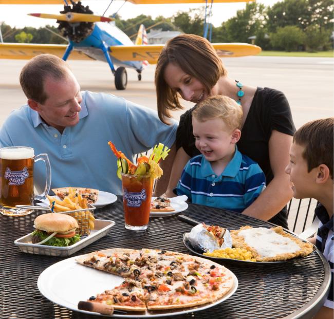 A family enjoys burgers and pizza on the patio at Stearman Field while a small prop plane is parked on the landing strip behind them