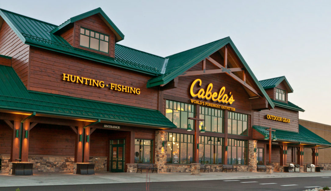 a storefront, it is a brown building with a hunter green roof and awning, there is yellow writing on the building that says "hunting", "fishing", "Cabela's World's Foremost Outfitter", and "Outdoor Gear".