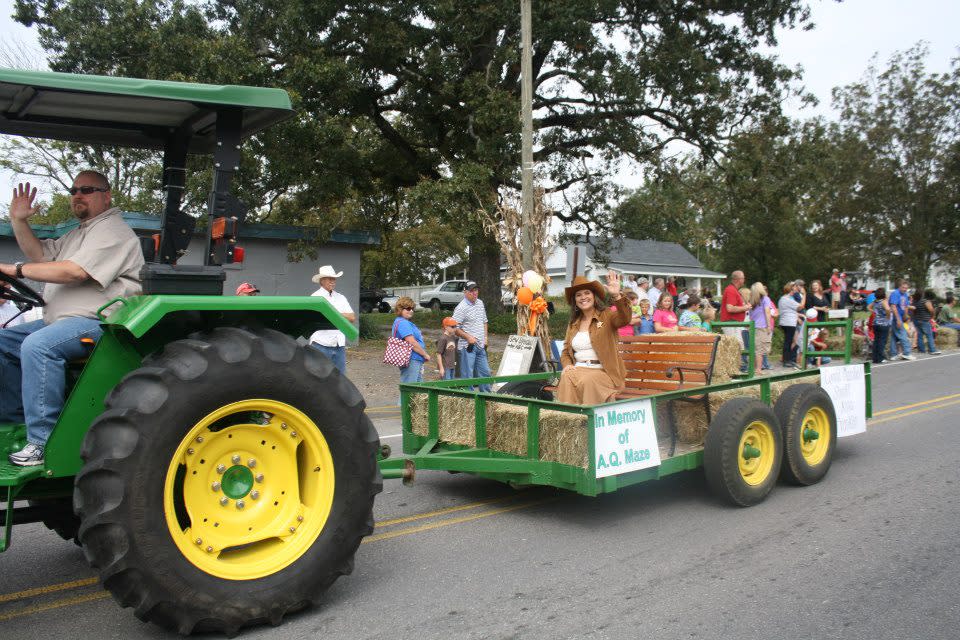 A man on a large green tractor pulls a trailer where a woman sits on hay bale waving to the crowd during Eva Frontier Days