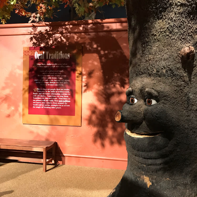 A tree with a face carving stands next to a sign that says "Oral Traditions" at EarlyWorks Children's Museum