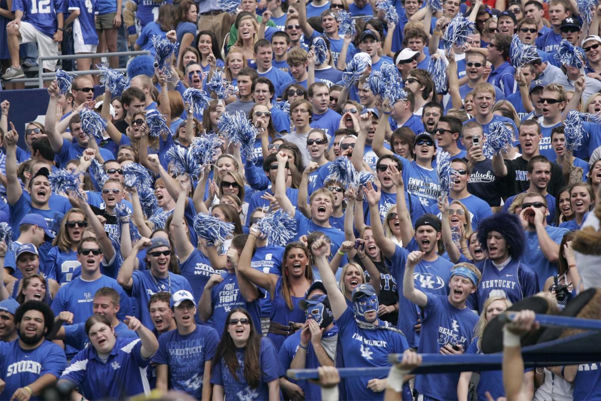 A huge crowd of excited fans all wearing University of Kentucky blue t-shirts cheers and waves blue and silver pom poms in the air.