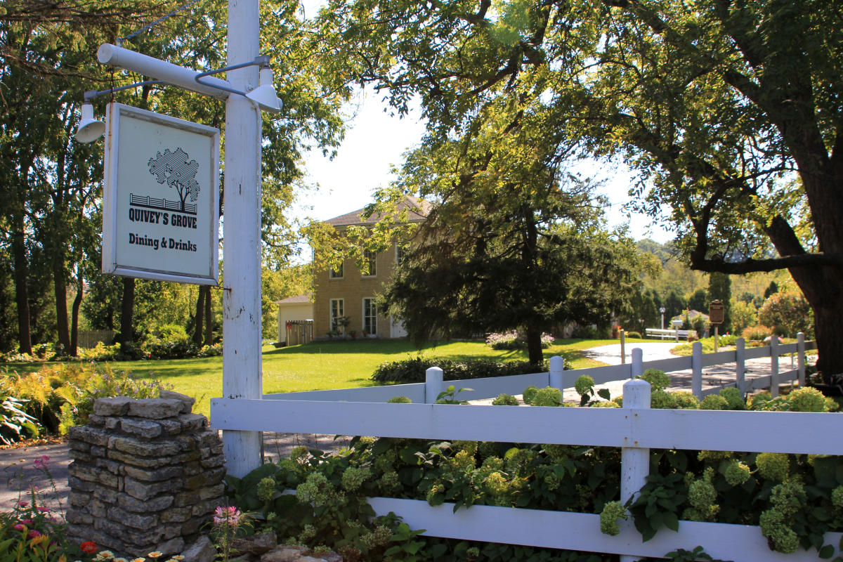 A sign that says "Quivey's Grove-Dining and Drinks" is at the entrance of a driveway. The sign is connected to a white, wooden fence, with a large, square brick house in the background.