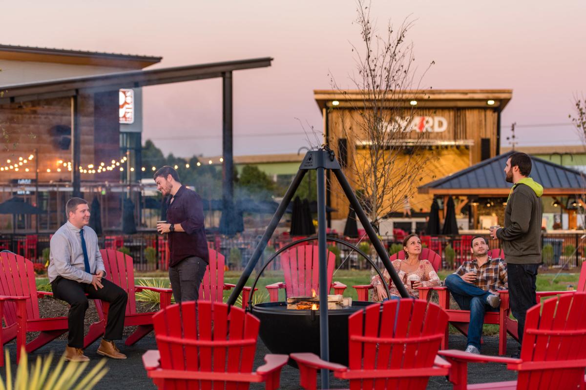 People sitting around a fire in red Adirondack chairs, in the back there is a building and an outdoor stage