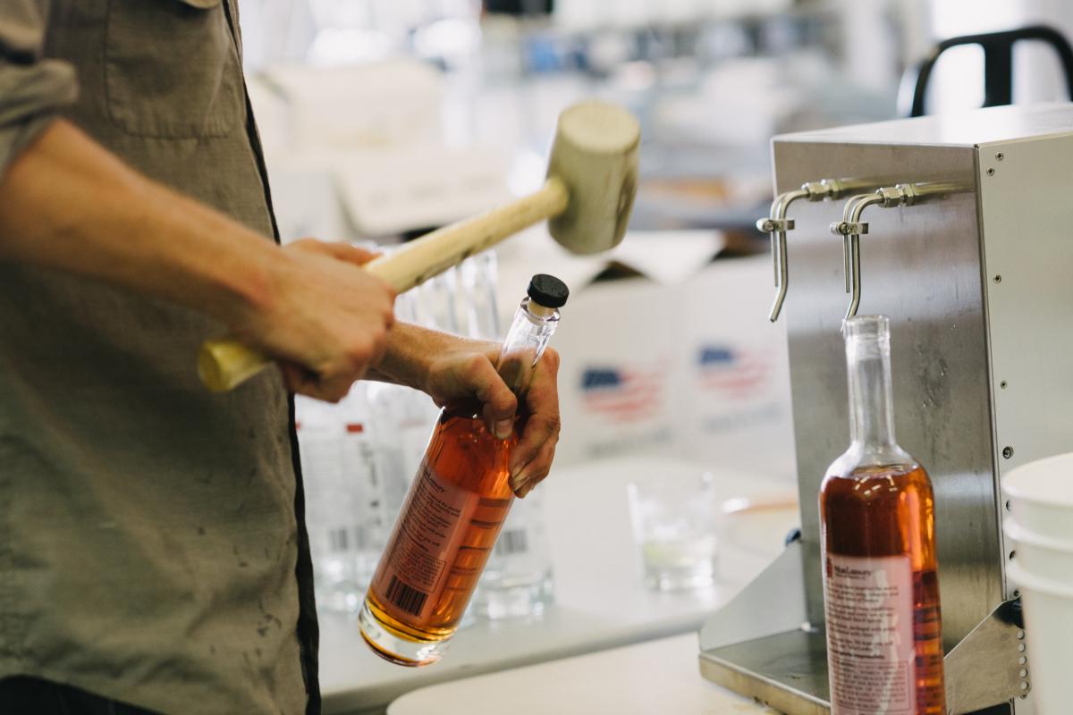 A person holding a mallet, tapping tops on bottles to seal them