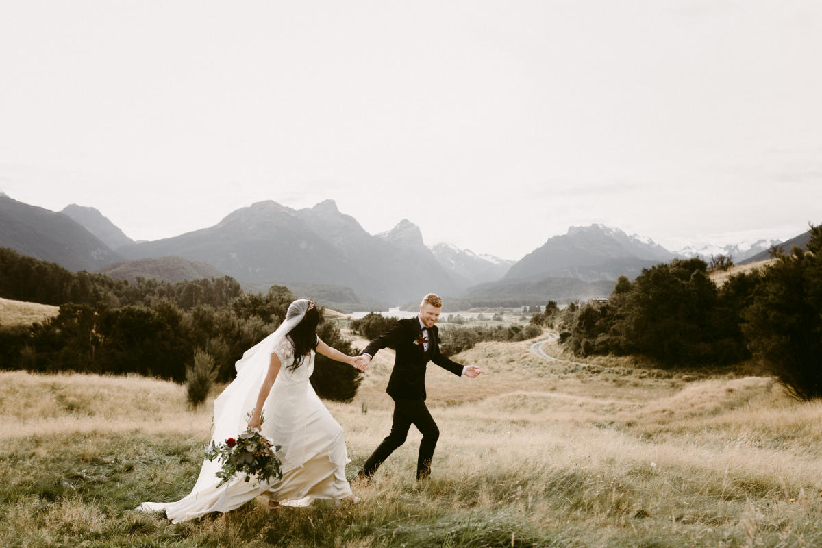 Married couple take wedding photos running through grassy field with mountain backdrop