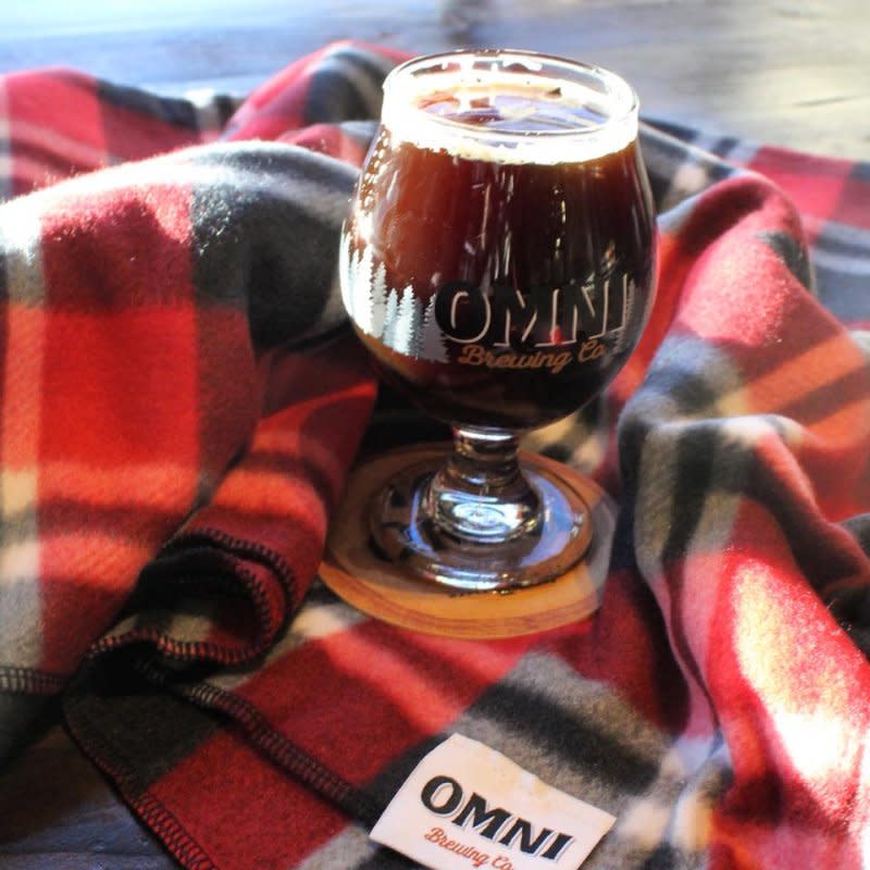 A glass of beer with the words "Omni Brewing Co" sits on a red and white flannel blanket