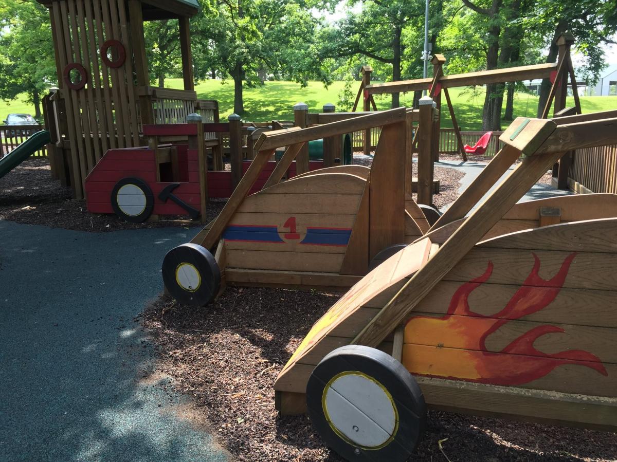 Three wooden, painted race cars for children to sit in. The closest car has flames on it. The second car as the number one on it and the third car has an ax on it.