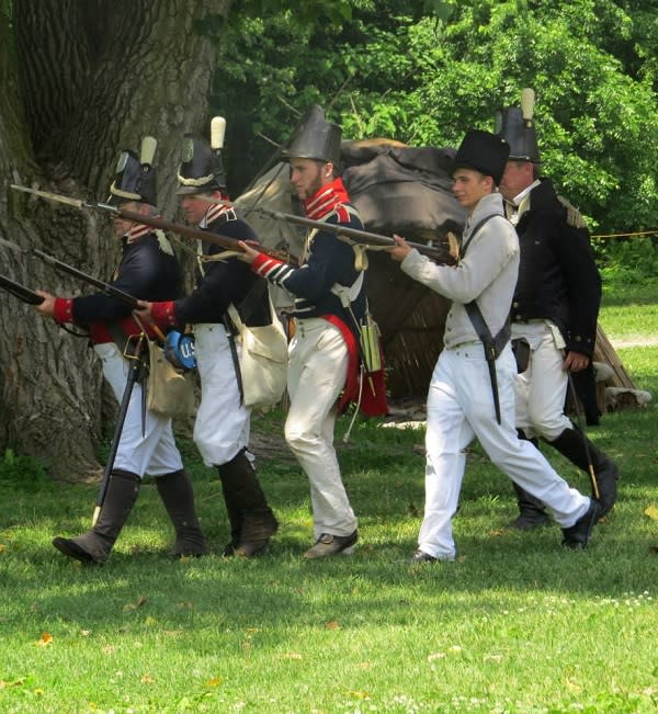 Notice the hats of these 1812 re-enactors? They're called Shakos. 1812 re-enactors hurry across the grass of the Fort
