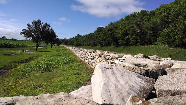 Limestone retaining wall along a curved road with a field, trees and blue skies.