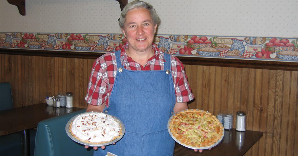 A woman in a red plaid shirt and overalls holds a pie in each hand at Village Inn Restaurant in Middlebury, Indiana