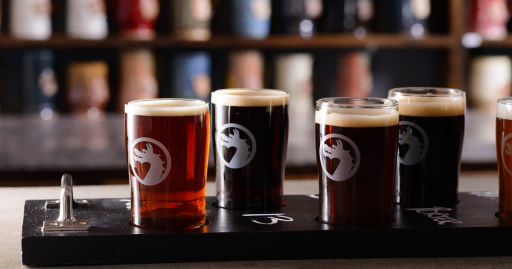 Five beers in glasses with a dragon logo sit on a wooden board on a bar top with out-of-focus taps in the background