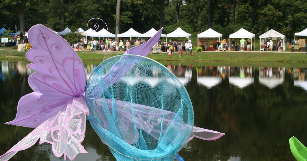 Close-up of a butterfly sculpture made of a butterfly net in front of a pond with festival tents set up on the other side of the water