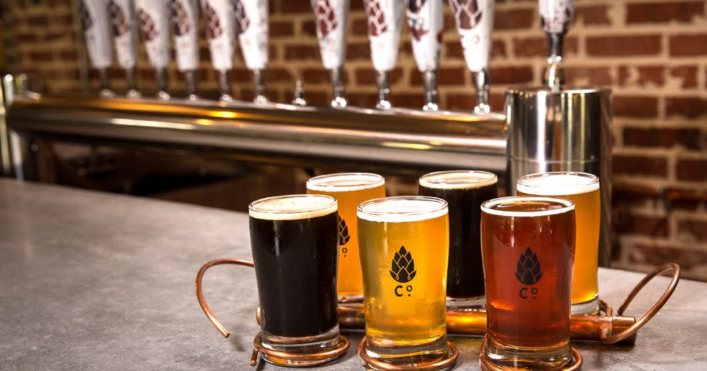 A flight of six beers in glasses marked "Co." sit on a bar in front of a row of white taps