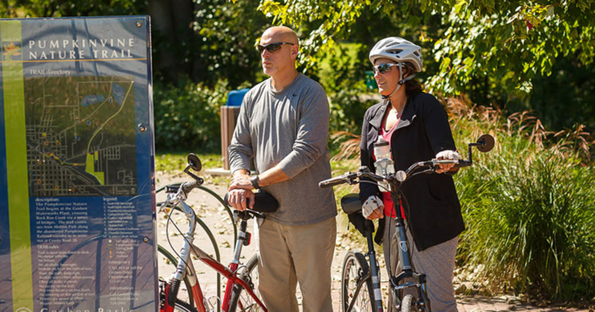 A man and woman stand next to their bicycles as they look at a sign with a map that says "Pumpkinvine Nature Trail"