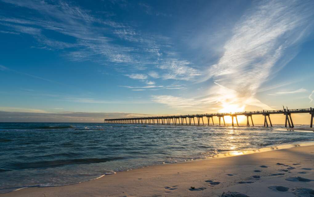 footprints along the shoreline next to a long dock by the ocean waters of pensacola beach at sunset