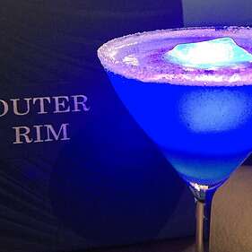 a salted rim dark blue cocktail with a glowing ice cube placed next to a bar menu that says outer rim
