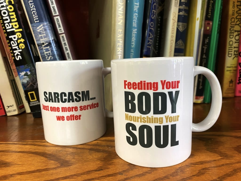 Two coffee mugs in front of a shelf of books. One reads "Sarcasm...just another service we offer" and the other "Feed your body. Nourishing your soul."