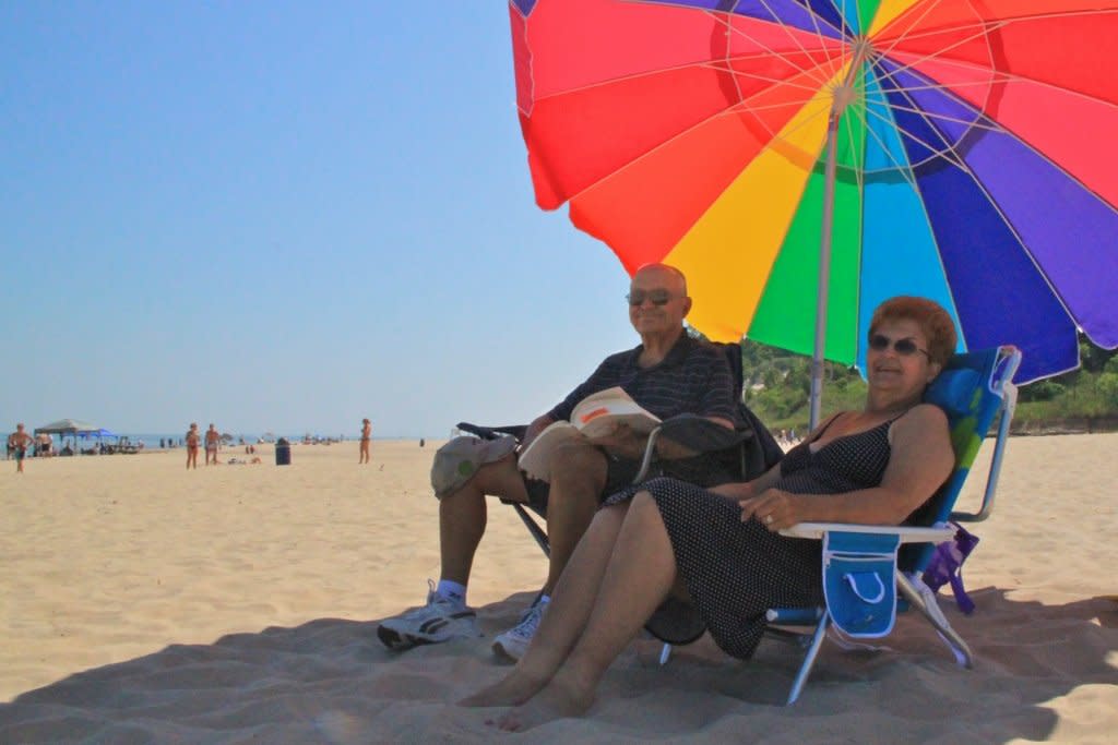 An older man and women sitting on the beach under a colorful umbrella