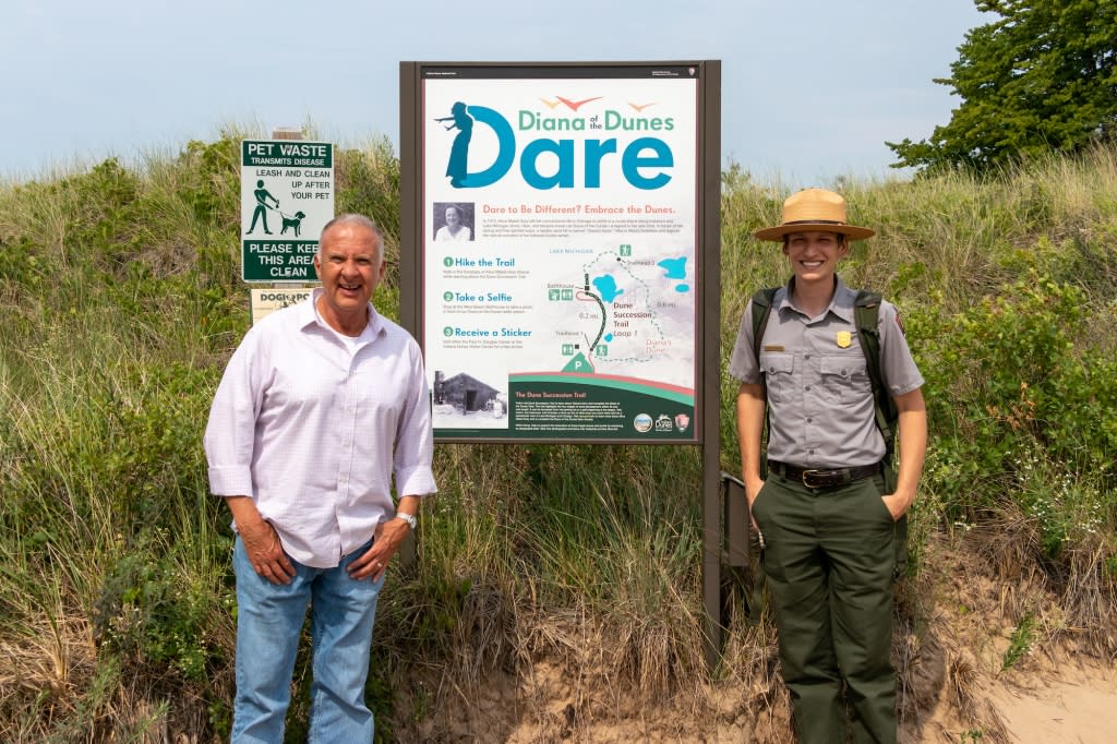 A man in a button down shirt and a park ranger stand in front of a trail sign outdoors.