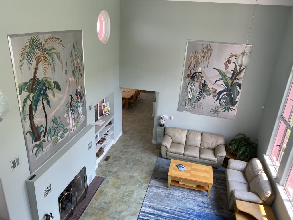 Interior shot of a house. Two tropical paintings hang on walls above a tan couch and a fireplace. 