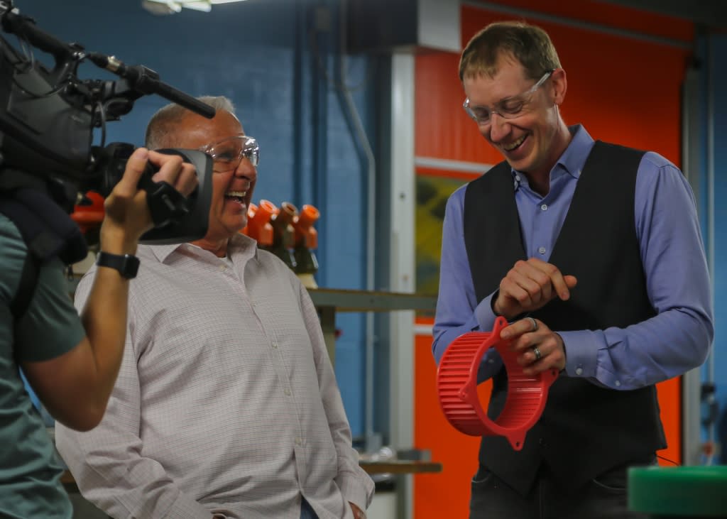 Two men wearing safety goggles look at a red object and discuss in front of a camera. 