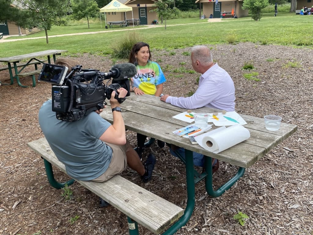 A gray-haired man sits at a picnic table with a dark-haired woman wearing a tie-dye tshirt. A cameraman sits across from them, filming.