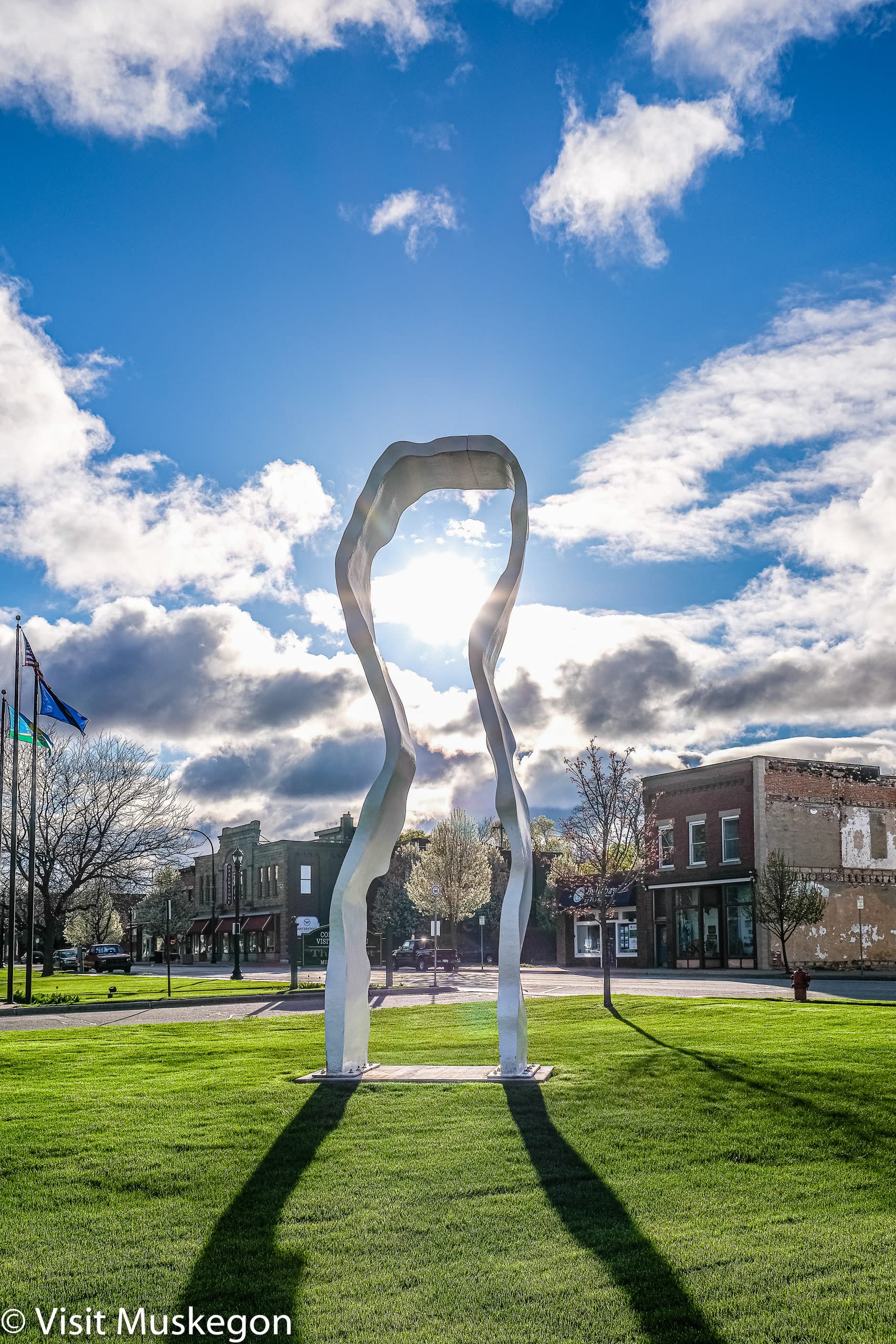 white sculpture that resembles arched noodle stands on green grass. Blue sky, white clouds and brick building are behind. flags can be seen to the left. 