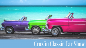 purple, green and pink calsic cars lined up in fron to lake promoting Cruz’in Classic Car Show