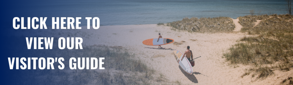 2 men carry surfboards at their sides as they walk over sandy path leading to lake michigan. text on left of image says click here to view our visitors guide