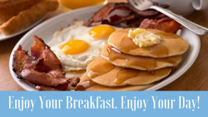 rectangular photo of scrambled eggs, bacon and biscuit on whte plate. pancakes visible in front right corner. fork leans on another plate in background. text in light blue bar says enjoy your breakfast enjoy your day.