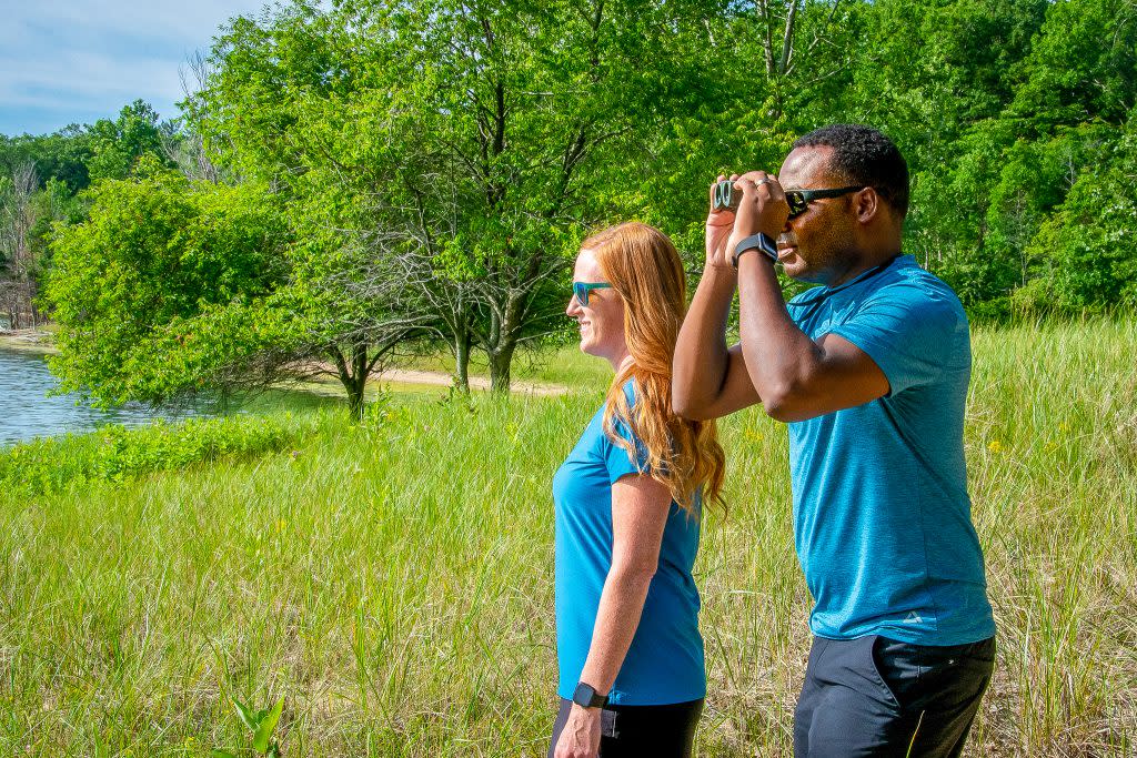Young couple stand in field with green trees in distance. Male uses binoculars to look out over lake which can be seen at edge of photo