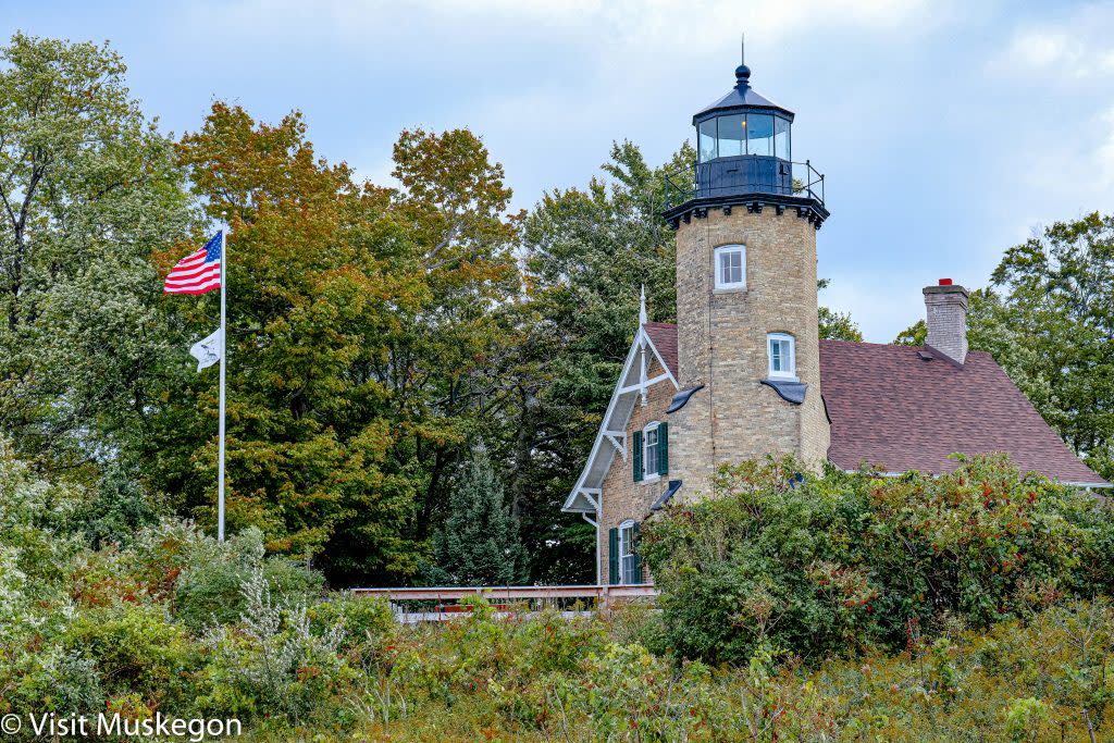 brick light house and tower sits on grassy hill surrounded by green trees. flags fly on pole in front of it. black iron clad light tops tower.
