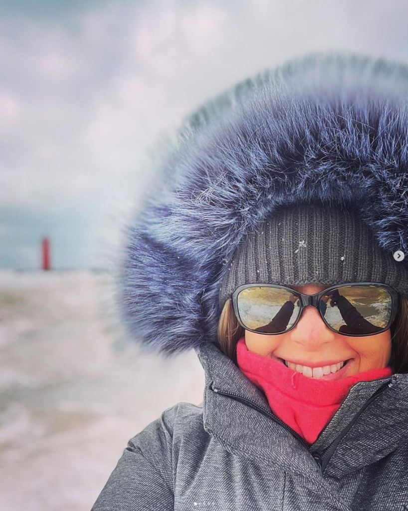 smiling woman's face is framed by fur trimmed coat hood as she stands in front of snow covered beach with lake michigan light tower in background